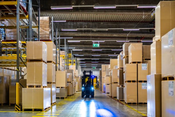 Warehouse worker driving forklift in aisle illuminated with headlights, wide shot